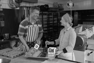 Zwei Personen lachen in der Backstube zwischen Brownies, Cookies und Backblechen.