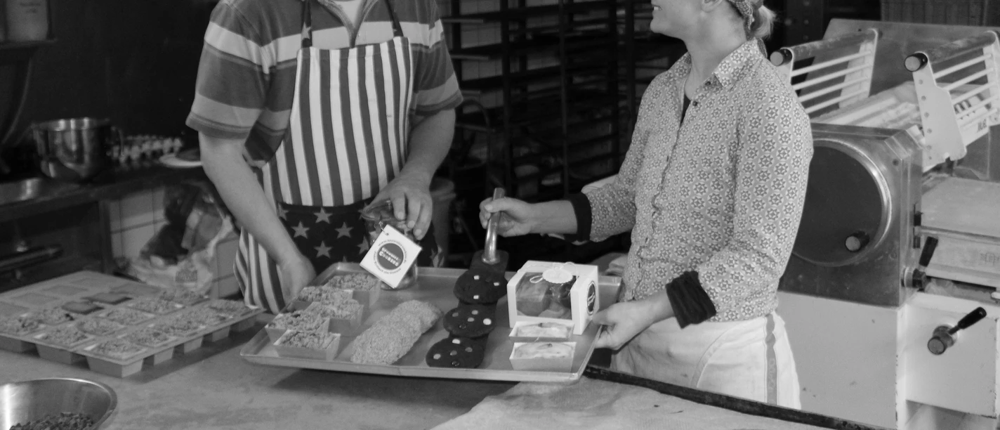 Zwei Personen lachen in der Backstube zwischen Brownies, Cookies und Backblechen.