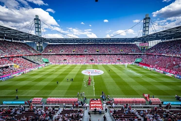 A packed RheinEnergieSTADION in Cologne, with a soccer field and fans under a blue sky