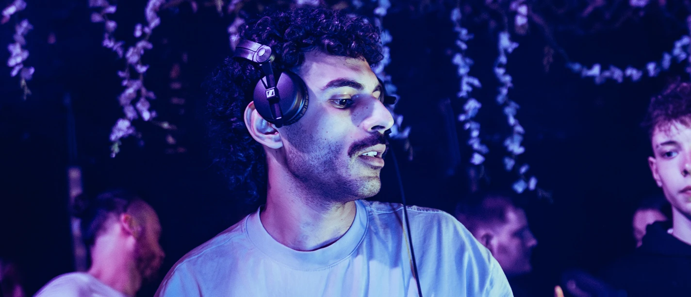 DJ an einem Mischpult in blau beleuchteter Umgebung mit hängenden Blumen.