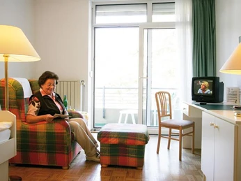Zimmer in der Klinik Waldfrieden Ein gemütlich eingerichtetes Patientenzimmer mit einer Frau, die auf einem Sessel sitzt und liest.