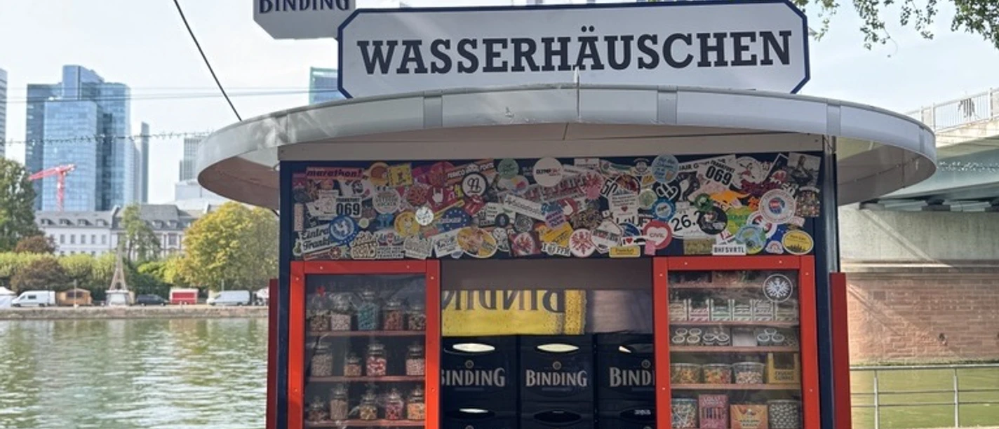 Bunt beklebtes Wasserhäuschen am Main mit Binding-Schild und Frankfurter Skyline im Hintergrund