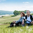 Kurze Rast auf dem Gesundheits - & Fitnessparcours Ein Paar sitzt entspannend auf einer Wiese in der Natur und genießt die Aussicht auf Hügel.
