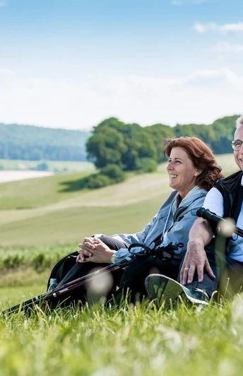 Kurze Rast auf dem Gesundheits - & Fitnessparcours Ein Paar sitzt entspannend auf einer Wiese in der Natur und genießt die Aussicht auf Hügel.