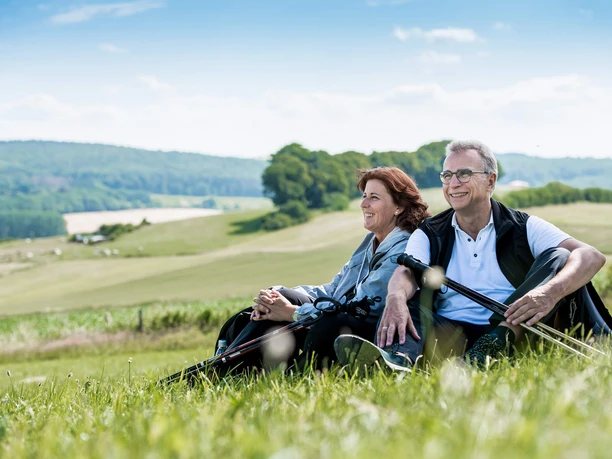 Kurze Rast auf dem Gesundheits - & Fitnessparcours Ein Paar sitzt entspannend auf einer Wiese in der Natur und genießt die Aussicht auf Hügel.
