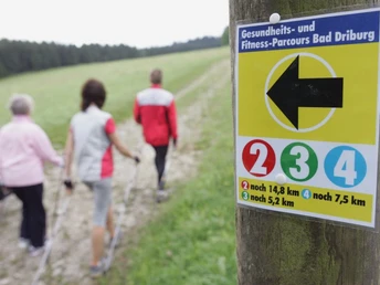 Zwei Frauen und ein Mann beim Nordic Walking entlang eines beschilderten Fitness-Parcours im Grünen.