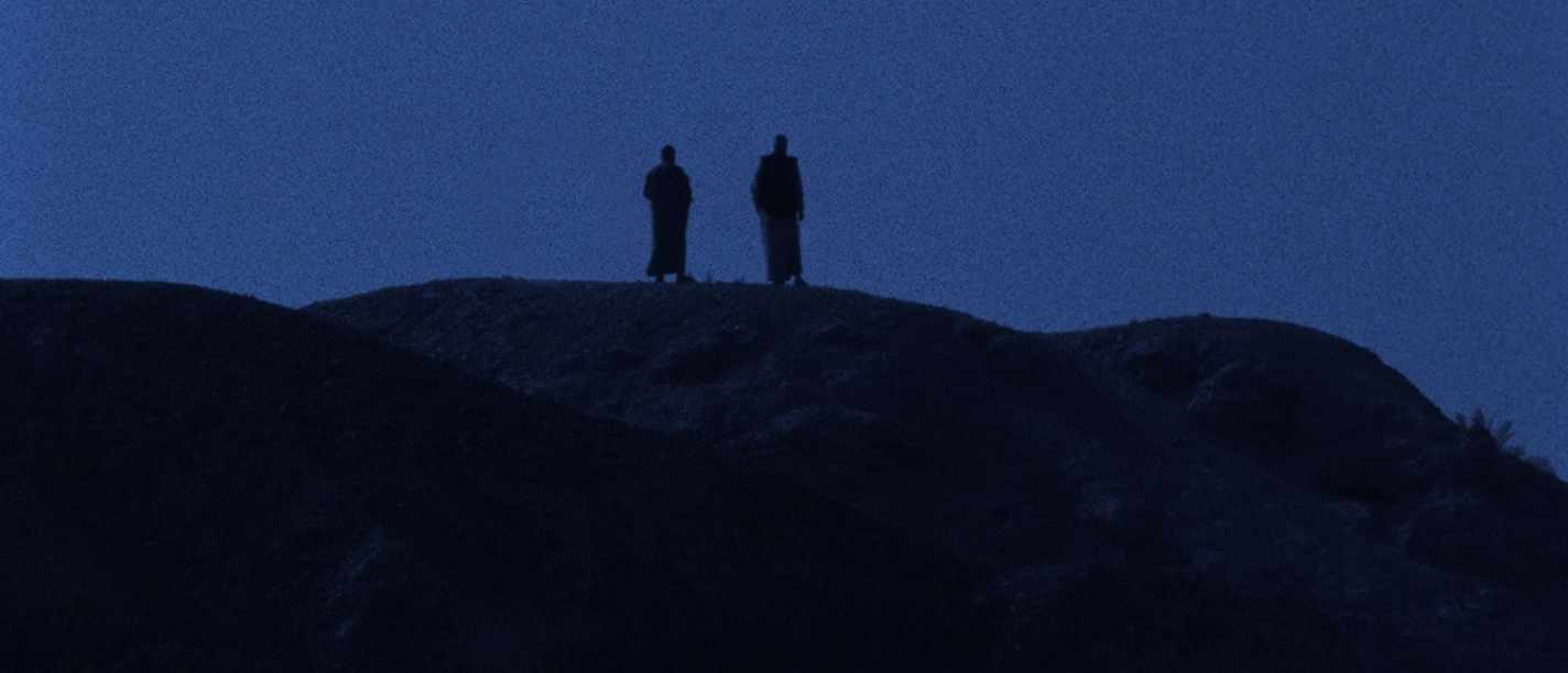 Zwei Personen stehen in weiter Ferne auf einer Felslandschaft bei einsetzender Dämmerung.
