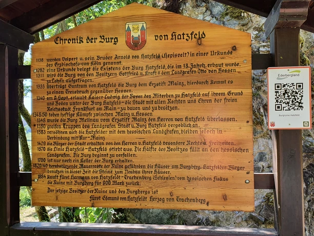 Hier ist eine Holz-Infotafel zu sehen, auf der die Geschichte der Burgruine zu erlesen ist.