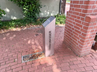 A metal drinking fountain on red cobblestones next to a brick wall.