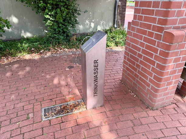 Metallischer Trinkwasserbrunnen auf rotem Pflaster neben einer Backsteinmauer.