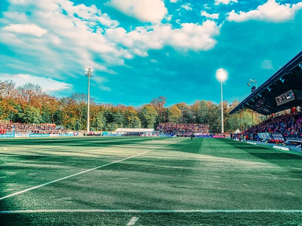 Fußballfeld im Stadion Höhenberg mit Tribüne, Flutlicht und Zuschauern unter blauem Himmel