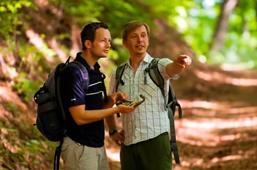 Unterwegs auf dem GPS-Erlebnispfad "Erlesene Natur - Waldwelten" Zwei Männer wandern auf einem Waldpfad unter grünen Blättern, einer zeigt lächelnd in die Ferne.