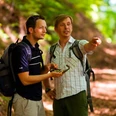 Unterwegs auf dem GPS-Erlebnispfad "Erlesene Natur - Waldwelten" Zwei Männer wandern auf einem Waldpfad unter grünen Blättern, einer zeigt lächelnd in die Ferne.