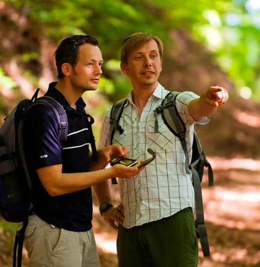 Unterwegs auf dem GPS-Erlebnispfad "Erlesene Natur - Waldwelten" Zwei Männer wandern auf einem Waldpfad unter grünen Blättern, einer zeigt lächelnd in die Ferne.