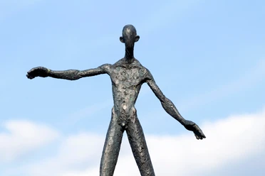 Dunkle Skulptur einer balancierenden Figur mit ausgestreckten Armen vor blauem Himmel