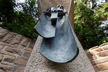Bronzerelief Klausner Heinrich an Steinstele zwischen Mauern und Bäumen, Ansicht von unten nach oben