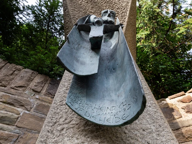 Bronzerelief Klausner Heinrich an Steinstele zwischen Mauern und Bäumen, Ansicht von unten nach oben