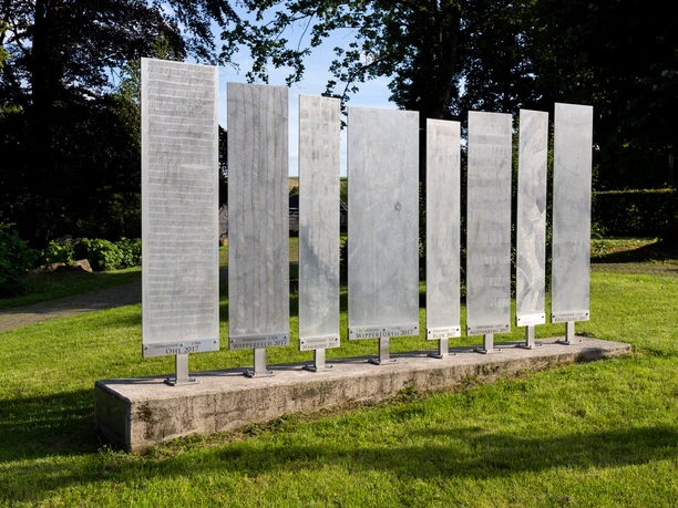 Metallene Ort-und-Zeit-Tafeln von Daphna Koll auf einer Wiese mit Bäumen im Hintergrund
