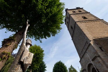 Bronzefigur „Der Prophet“ mit erhobenem Arm vor einem Kirchturm und Bäumen in Wipperfürth