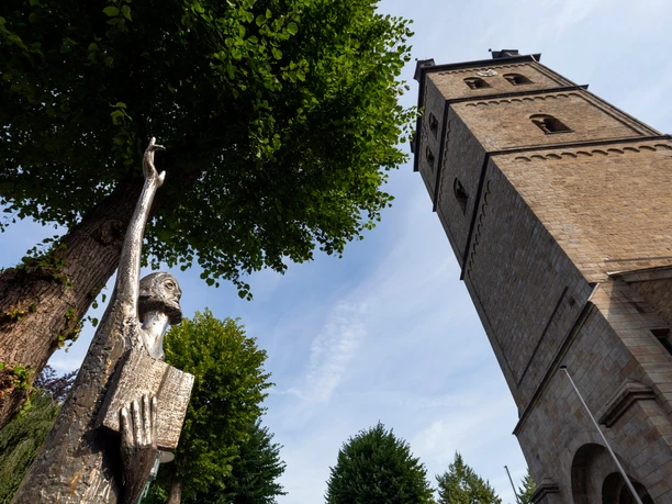 Bronzefigur „Der Prophet“ mit erhobenem Arm vor einem Kirchturm und Bäumen in Wipperfürth
