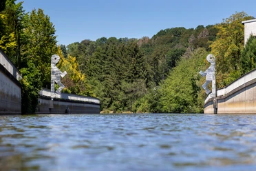 Zwei silberne Figuren auf Brückenpfeilern über der Wupper vor grünen Bäumen