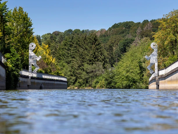 Zwei silberne Figuren auf Brückenpfeilern über der Wupper vor grünen Bäumen