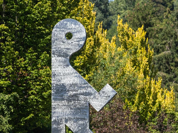 Metallskulptur einer schreitenden Figur an der Wupper vor grünen Bäumen
