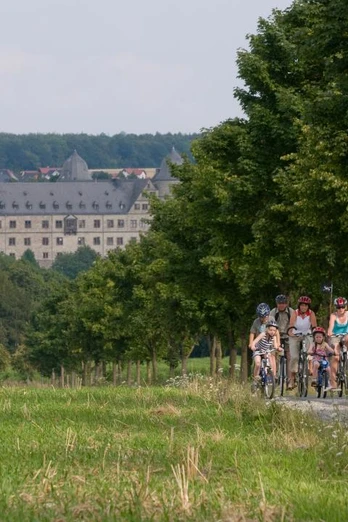 Familie auf Fahrradtour nahe einer historischen Burg, umgeben von grüner Natur unter blauem Himmel.