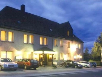 Haus Engemann Mehrstöckiges Gebäude mit beleuchteten Fenstern am Abend, Parkplätze und Fahrzeuge im Vordergrund.