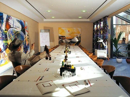 Konferenzraum im Sporthotel Wiedenbrück Ein Konferenzraum mit langer Tafel, moderne Kunst an den Wänden, eine Person sitzt entspannt am Tisch.