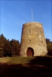 Kumsttonne_Herbstansicht Eine alte, steinerne Windmühle steht vor einem Hintergrund aus herbstlichen Bäumen unter klarem Himmel.