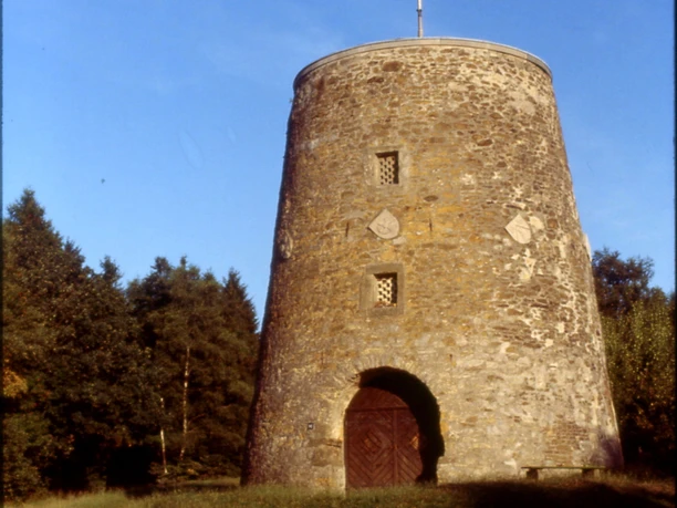 Kumsttonne_Herbstansicht Eine alte, steinerne Windmühle steht vor einem Hintergrund aus herbstlichen Bäumen unter klarem Himmel.