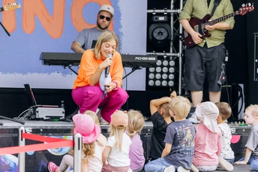 Singer Nina is crouched in front of the children with a microphone; the band is playing on stage in the background