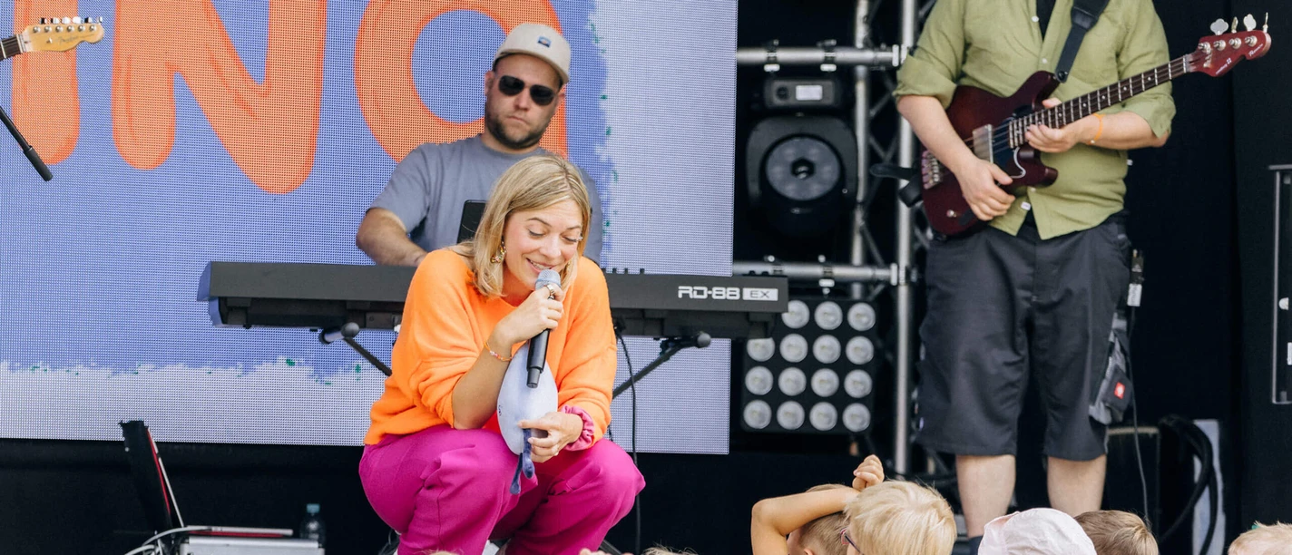Sängerin Nina hockt mit Mikrofon vor Kindern, Band spielt im Hintergrund auf der Bühne