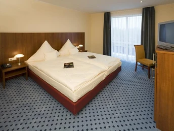 Helles Hotelzimmer mit zwei großen Betten, Gästeordner auf den Betten, Schreibtisch, Stuhl und Fernseher.