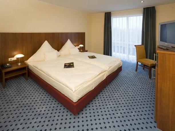 Zimmer im Airport Hotel Paderborn Helles Hotelzimmer mit zwei großen Betten, Gästeordner auf den Betten, Schreibtisch, Stuhl und Fernseher.