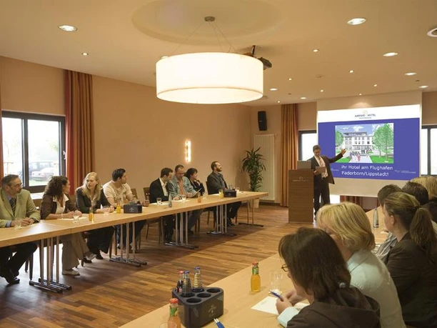 Seminarraum im Airport Hotel Paderborn Seminarraum mit Teilnehmern an Tischen, Vortragender an der Präsentationsfläche, moderne Ausstattung.