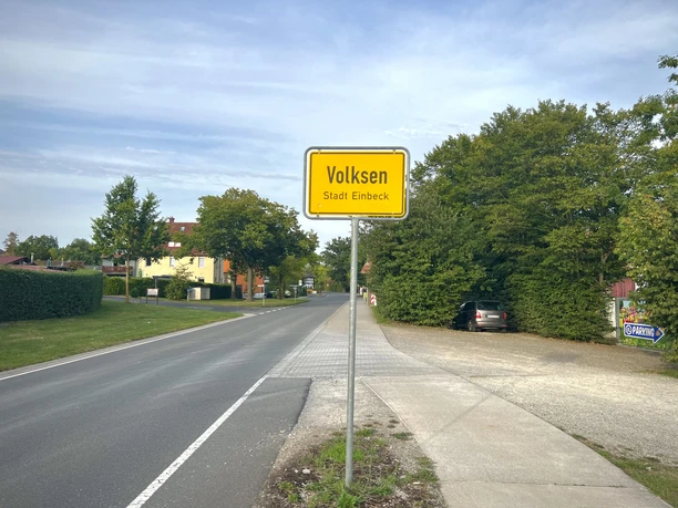 Gelbes Ortsschild mit der Aufschrift Volksen Stadt Einbeck am Straßenrand vor Bäumen.