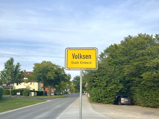 Gelbes Ortsschild von Volksen Stadt Einbeck am Rand einer Straße mit Bäumen und blauem Himmel.