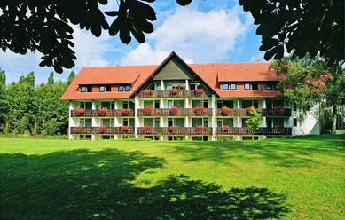 Außenansicht auf das HolsingVital HolsingVital Außenansicht mit rotem Dach, Balkonen und gepflegtem Rasen, eingefasst von Bäumen.