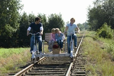 Draisine Familie mit zwei Kindern fährt auf einer Draisine durch eine grüne, von Bäumen gesäumte Landschaft.