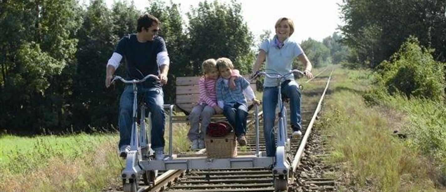 Draisine Familie mit zwei Kindern fährt auf einer Draisine durch eine grüne, von Bäumen gesäumte Landschaft.