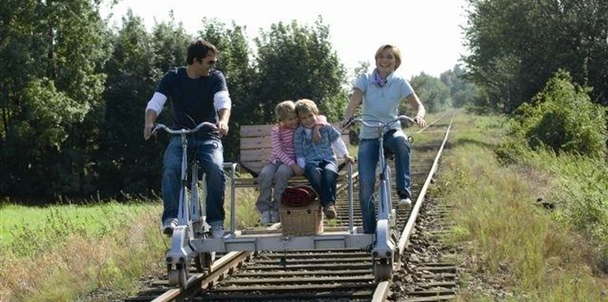 Draisine Familie mit zwei Kindern fährt auf einer Draisine durch eine grüne, von Bäumen gesäumte Landschaft.