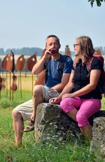 Zwei Personen sitzen auf Steinen und unterhalten sich, im Hintergrund stehen rostrote Skulpturen.