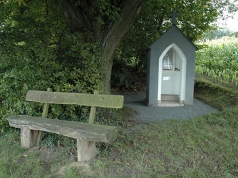 Eine kleine Kapelle mit Kreuz steht unter einem Baum, daneben eine hölzerne Parkbank.