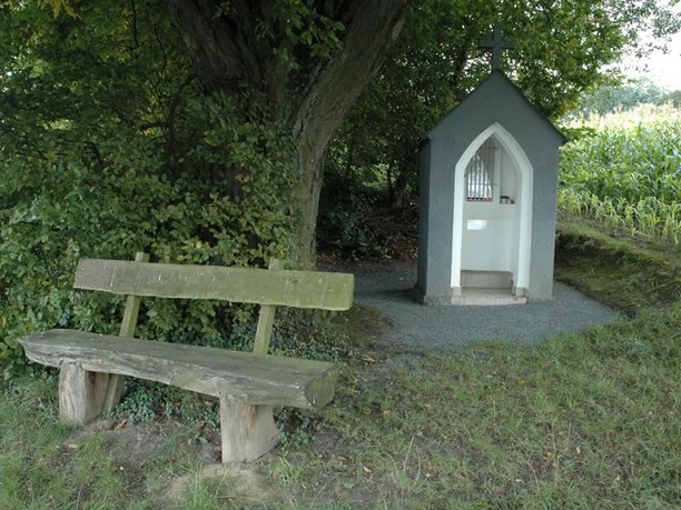 Eine kleine Kapelle mit Kreuz steht unter einem Baum, daneben eine hölzerne Parkbank.