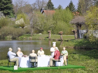 "Alltagsmenschen" am Emssee Eine Gruppe von acht lebensgroßen Skulpturen sitzt an einem Tisch im Freien am Ufer eines Teiches.