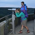 Eine Frau blickt durch ein Fernrohr auf einer Aussichtsplattform, umgeben von Waldlandschaft.