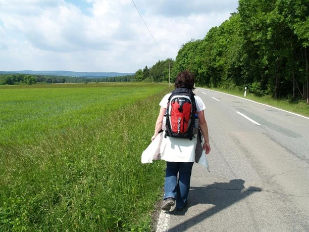 Auf der Emder Höhe Person mit Rucksack wandert im Sonnenschein auf einer Landstraße neben üppig grünen Wiesen.