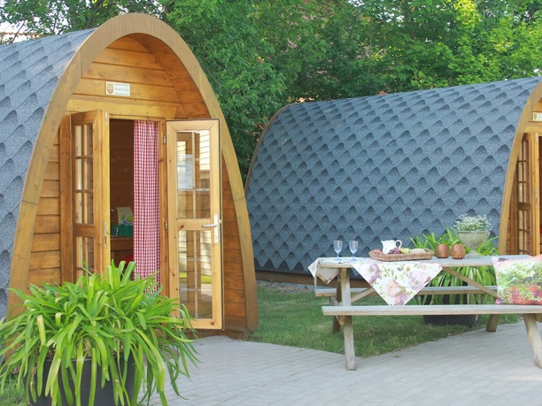 Holz-Camping-Pods für je vier Personen, umgeben von grüner Natur, mit Picknicktisch ausgestattet.